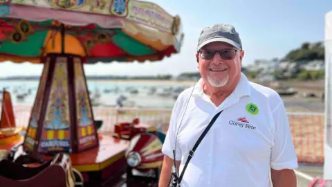 Lester Richardson is pictured in a white polo shirt. Mr Richardson is wearing a cap and sunglasses and he is smiling widely. There is a carousel behind him and a beach is also visible in the background.