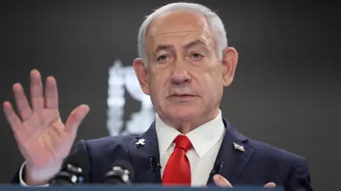 Reuters Benjamin Netanyahu, who has grey combed over hair, and wears a navy suit with and Israel badge and a yellow ribbon badge - which is a symbol calling for the release of hostages currently held by Hamas - on his lapel, holds a hand up as he delivers a press conference