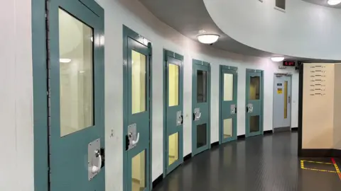 A row of custody suites along a curved wall. The reinforced doors to the custody suites are blue and have lights hanging above them.