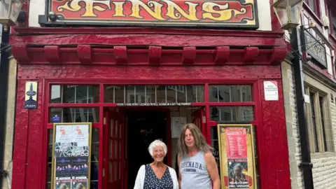 A woman white short white hair, wearing a dark blue dress with a pattern of white dots and a white cardigan stands in the entranceway to a pub which has the sign 'Finns' over the door, which is painted dark red. To her left is a man with long brown hair past his shoulders, wearing a sleeveless grey t-shirt with a logo across the chest and blue jeans.