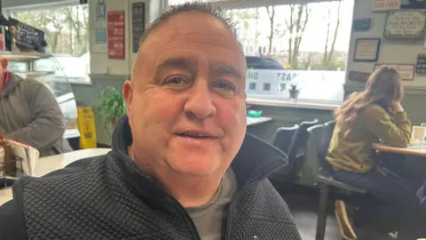 Kevin seated at a table inside a busy café. He is wearing a dark zip‑up jacket over a T‑shirt. Behind him, other customers are seated at tables. One person is sitting at a nearby table on the left with condiments and dishes in front of them. Another customer sits further back on the right, facing sideways toward a window. The café walls are decorated with numerous signs and framed items, and large windows let in daylight, showing trees and parked vehicles outside.