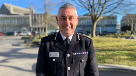 A police officer in uniform smiling at the camera. He is standing outdoors in front of a grass bank and building. Trees are in the background. The sky is blue. 
