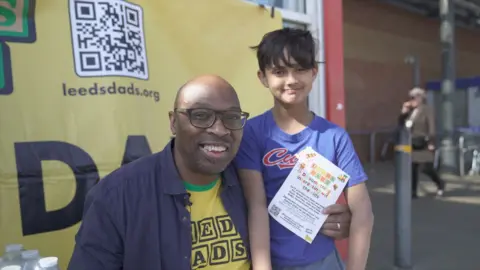 Hannah Karpel / BBC Man crouched down with his arm around his young son in front of a yellow sign that reads Leeds Dads. He hold a flyer advertising the group. Both smile into the camera.