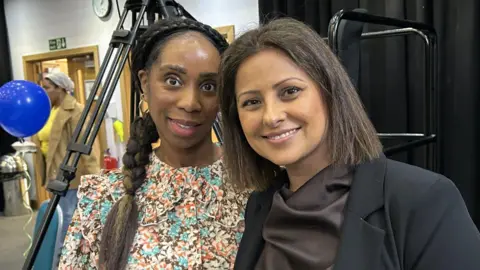 A woman with long dark hair and a floral top standing next to a woman with dark hair, a dark jacket and dark brown top in a room with a black tripod behind them