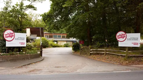 The White Lodge facility in Chertsey, Surrey. Two white signs welcoming people into the facility sit either side of the wooden gates, and the facility building can be seen in the image. 