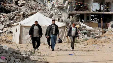 Reuters Palestinians walk among rubble in Rafah, with a tent and a bombed out building in the background (March 2025)