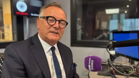 A man wearing a grey suit jacket, white shirt and dark blue tie with white spots on. He is sat in a chair in a radio studio looking into the camera. In front of him is a purple microphone with the words BBC RADIO SUSSEX written on it in white lettering