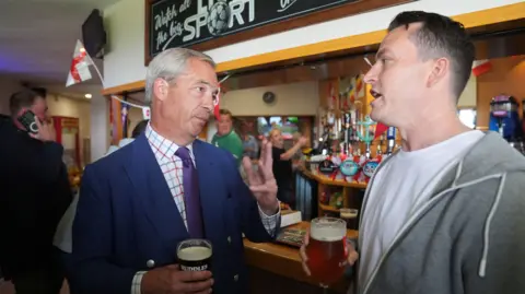 PA Media Nigel Farage in a suit with a pint in his hand. A man in his late thirties (McMurdock) is wearing a white T-shirt and grey hoodie and also holding a pint.