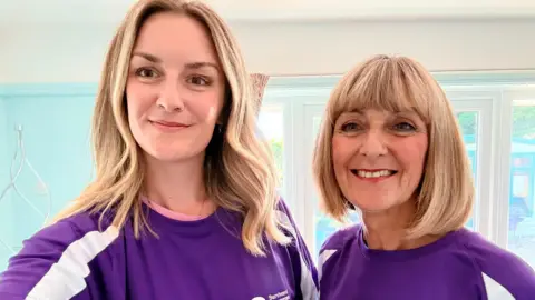 Alyson Noakes and her daughter Siobhan. The pair are wearing matching purple T-shirts and are stood in a house with a blue wall behind them. Alyson has shoulder-length blonde hair and her daughter as slightly longer, wavy blonde hair.