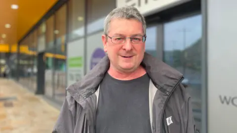 Josh Tranter/BBC A man with grey hair wearing glasses and a grey coat is looking at the camera. The building behind him is blurred.
