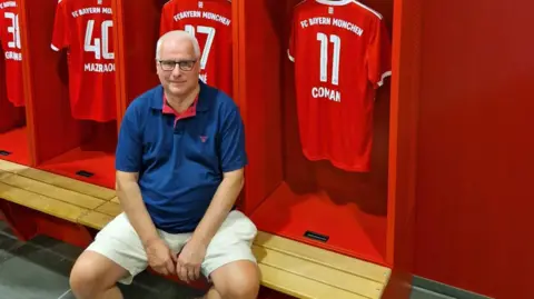 Mike Ash sitting on a bench in a changing room wearing a blue shirt and white shorts. Behind him are cubby holes with red football shirts hanging on hooks