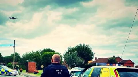 View of a police scene. In the centre the head and shoulders of a man in a police jacket, with a marked police car next to him on his right. To the left a police car is parked diagonally on the road next to a brick bus shelter. In the sky to the top left a drone can be seen.