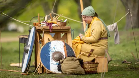 Great Ousekjarr Viking Society Woman in viking costume making clothes surrounded by Viking shields