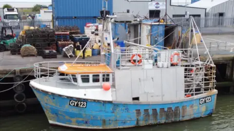 A blue and white fishing vessel at a mooring. There are a number of buoys on the side and two life rings. There are a number of buildings in the background.