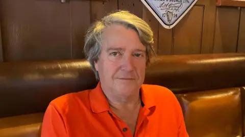 BBC The image shows a man wearing an orange top. He is looking directly at the camera. He is sitting on a brown padded seat and there is a wooden wall behind him.