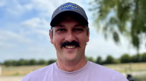 BBC/ Jess Lord A farmer, who wears a cap and a lilac t-shirt, smiles outdoors.