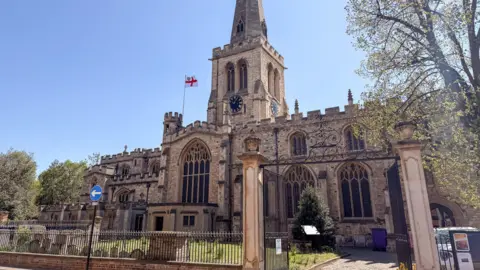 Anthony Davis A view of a church from the street, with railings around the perimeter of the grounds and cemetery and a large gate in the foreground. It is a sunny day with blue sky. The tower is prominent with its clock visible on two sides. There is a St George's flag flying on the roof.