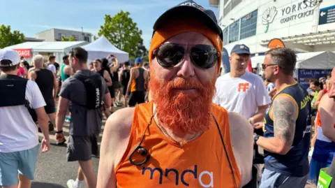 Katy Lambert/BBC An elderly man in an orange running vest. He has dyed his beard bright orange and wears an orange hat. He smiles at the camera.
