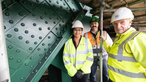 Newcastle City Council Councillor Juna Sathian, Newcastle City Council, Gareth Dawson, Esh Construction and councillor John McElroy, Gateshead Council are smiling as they stand by the newly-repainted in green sections of the Tyne Bridge. they are all wearing high visibility protective jackets and helmets. 