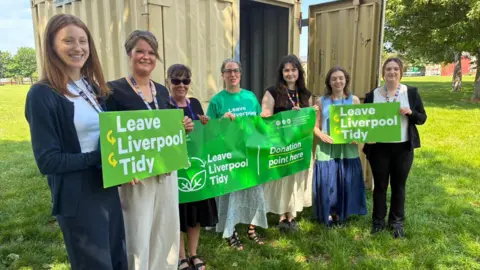 Seven women are standing in front of a large recycling container holding green banners with white writing which says Leave Liverpool Tidy