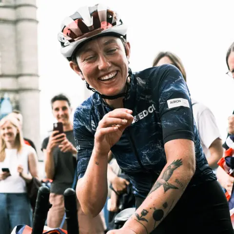 Rupert Hartley/Albion Cycling An exhauster woman wearing a red and white cycling helmet and dark blue top, smiles with her eyes almost closed as she sits on a bike. People behind her can be seen smiling and taking pictures with their phones.