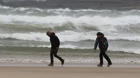 Two people bend into the wind as they walk along a beach with crashing waves behind them