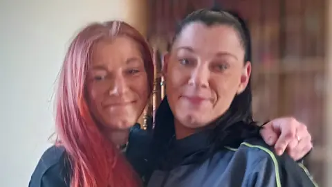 West Midlands Police Two women, one with red hair and another with black hair. The woman with red hair has her arm around the shoulder of the other woman. They are both smiling and the background is blurred. They are standing indoors in front of a wooden cupboard.