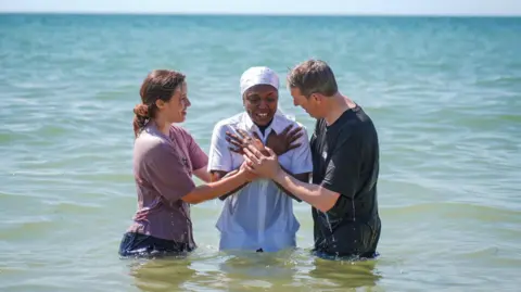Three people in the sea two either side of a person in all white who has been dunked in the water