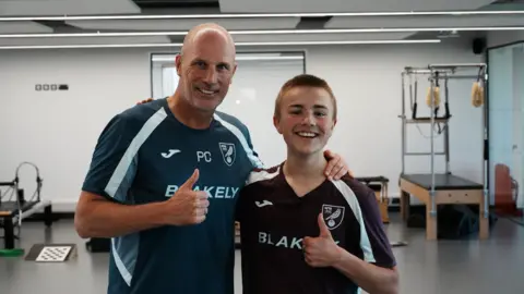 John Fairhall/BBC Norwich City's head coach Philippe Clement on the left in a green training top is pictured with Ranulph on the right in a maroon-coloured Norwich shirt. Both are giving a thumbs up and smiling at camera.