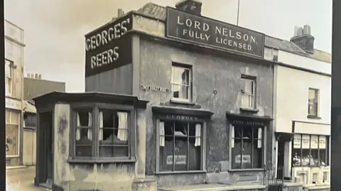 Geo Jones and Bros A black-and-white historic photo of a pub called The Lord Nelson - taken approximately 100 years ago. It has a three large windows facing the street and two smaller windows on the first floor. It occupies a corner position at the end of a row of shops, with a large sign for Georges' Beers across the corner wall.