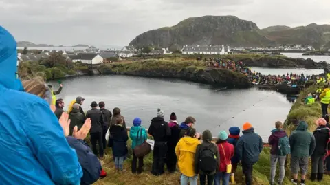 People watching the World Stone Skimming Championships in a flooded quarry, with white-walled houses of Easdale are in the backgroud