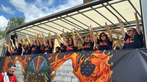 Women wearing black T-shirts are singing on board a lorry trailer. They have gold bracelets on and are holding their hands in the air.
