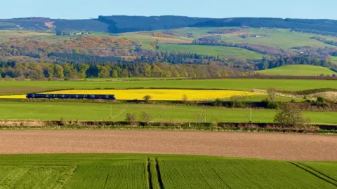 Lorna Donaldson A Passenger train travelling through patchwork farmland, with green and yellow fields stretching toward wooded hills under a clear blue sky.