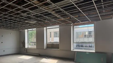 Annabel Amos/BBC Plaster-covered interior walls of a room,with the false ceiling removed and large square windows on the far side. The shadow from the windows is visible on the concrete floor.