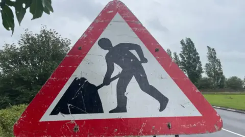 A triangular road work sign with a red border. Behind it is a road with a green field to the right and some trees and bushes.