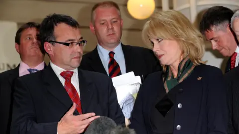 PA Phil Woolas, wearing a dark suit and red suit, talks with Joanna Lumley during a press conference