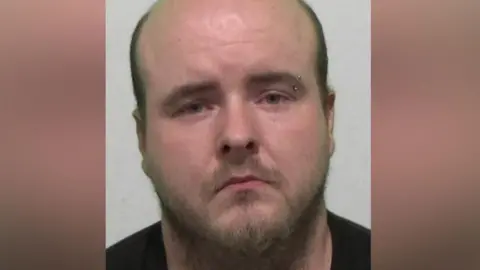 Mugshot of Rogers. He is bald with a scruffy brown beard and left eyebrow piercing.