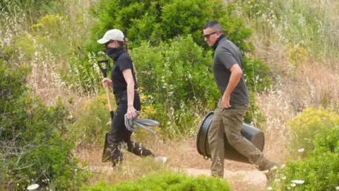 Officers, one carrying a space and the other a large plastic bucket, walk through the area near a derelict property to the west of Praia De Luz