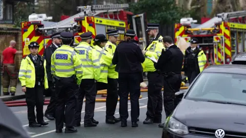 A group of police officers stand beside fire engines on a road