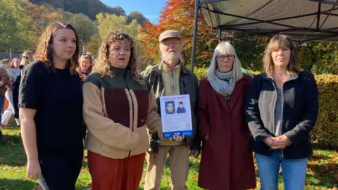 Two young women, an older man and two older women stand together in a park holding a poster appealing for information