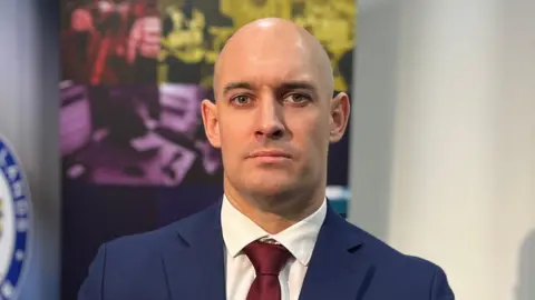 A man with a bald head in a blue suit with a red tie in a room with white walls and a blurred blue symbol on one wall