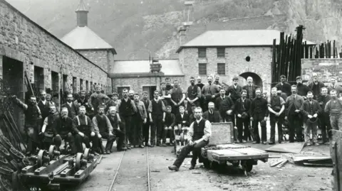 National Slate Museum Old black and white photograph of the quarry workshops with dozens of staff posing in the courtyard amid slate equipment