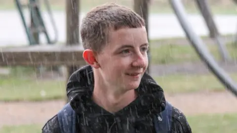 Keaton Lacey wearing a dark coat in the rain. He is looking away from the camera and there is grass and a path behind him.