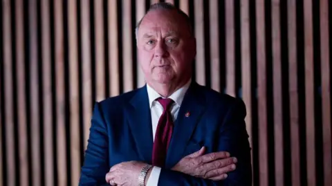 Getty Images Managing director of Mineral Resources Chris Ellison wearing blue suit, white shirt, and red tie with silver watch folding arms in front of wooden background.