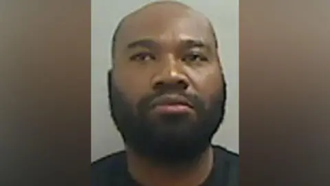 Cleveland Police Mugshot of Hammond. He has a thick black beard and a bald head and is unsmiling. 