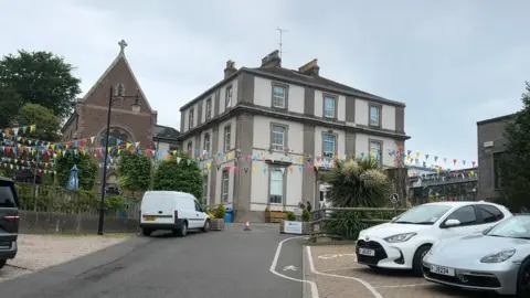 A school building is visible with a car park in the foreground. There is bunting and a church on the site. 