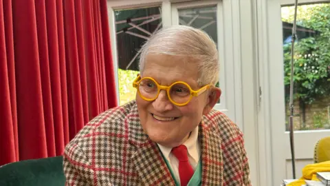 Jack Lupton David Hockney wearing a tweed suit with a red tie smiles as he wears a pair of bright yellow round glasses.