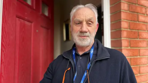 Roger Hewish looking into camera. He has white hair and a white beard, and is standing in front of a red front door which is ajar.
