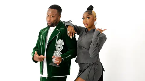 Donnie Sunshine/MOBOs Babatunde and Indiyah on a white background looking at the camera. Babatunde on the left is wearing a white tshirt under a green blazer, holding a silver award which spells out "MOBO". Indiyah on the right has her right arm over his shoulder, and is wearing a grey outfit with silver earrings. Her left hand is doing a "peace" sign.