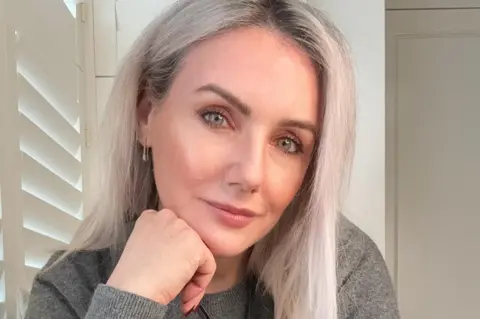 Sali Hughes A woman sits near a window with wooden blinds, resting her chin on her hand. She's smiling slightly, her pale blue eyes are piercing. She has grey hair cut into a mid-length style and wears a grey sweater.